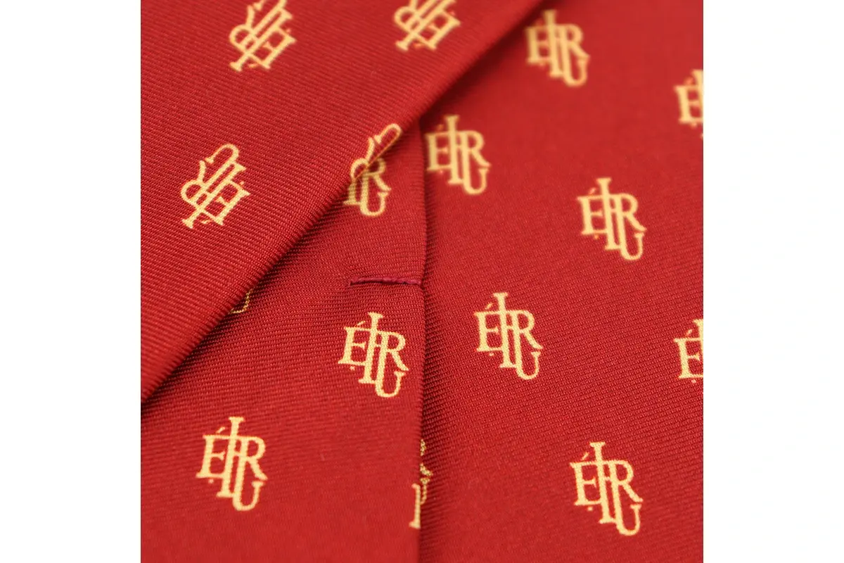 Gold letter monograms repeated across red woven tie fabric.