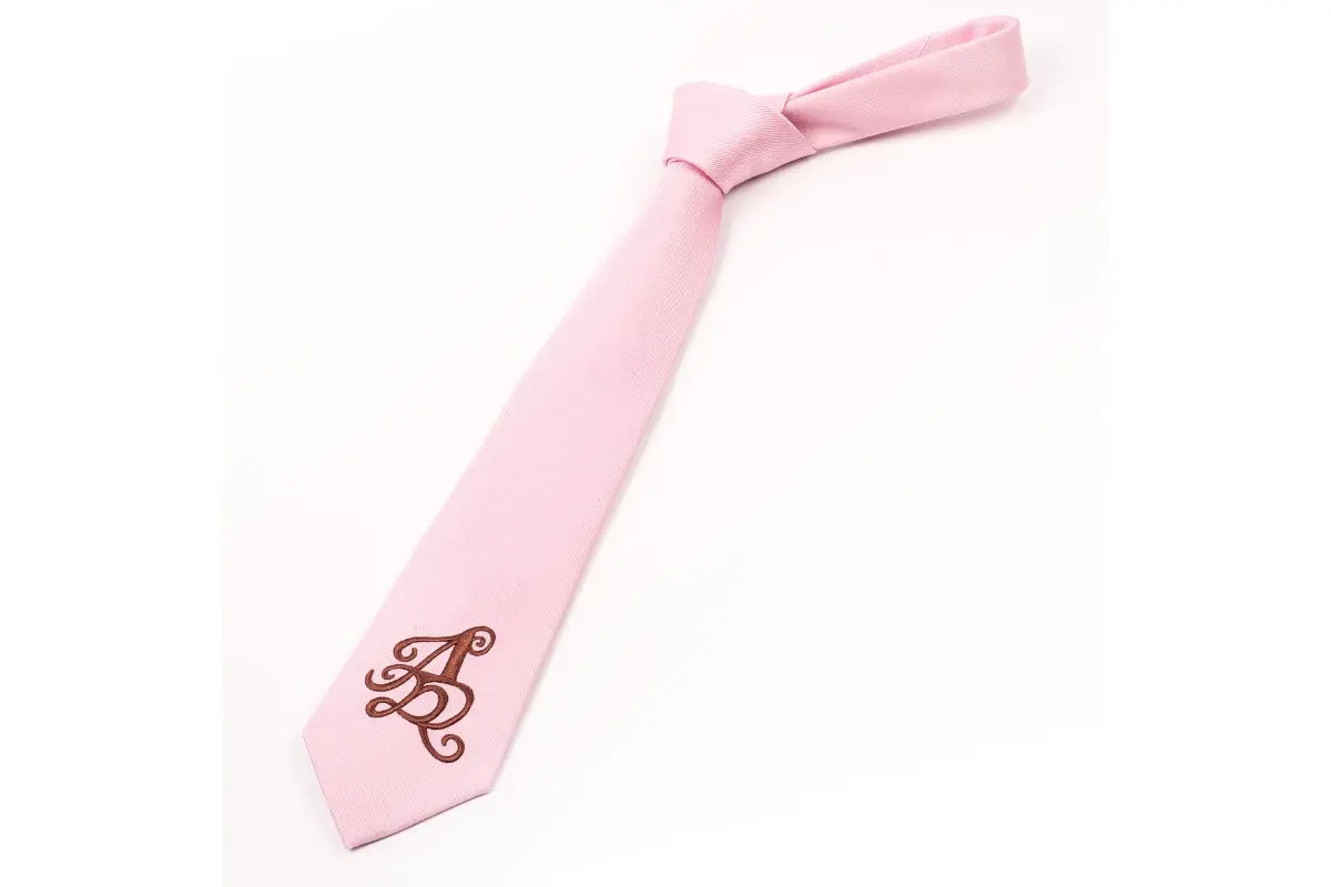Pink knotted tie with a burgundy monogram placed near the blade tip.